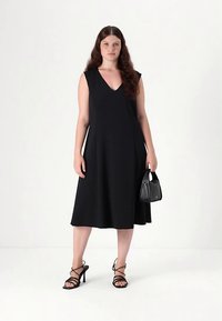 Svart ärmlös klänning med V-ringning, luftig A-linje silhuett, slätt tyg, kombinerad med svarta remhäskor och en liten handväska.