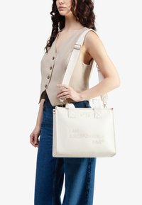 Borsa a tracolla bianca con texture e testo impresso. Dotata di doppie maniglie, tracolla regolabile e forma quadrata. Progettata con accessori hardware minimi.