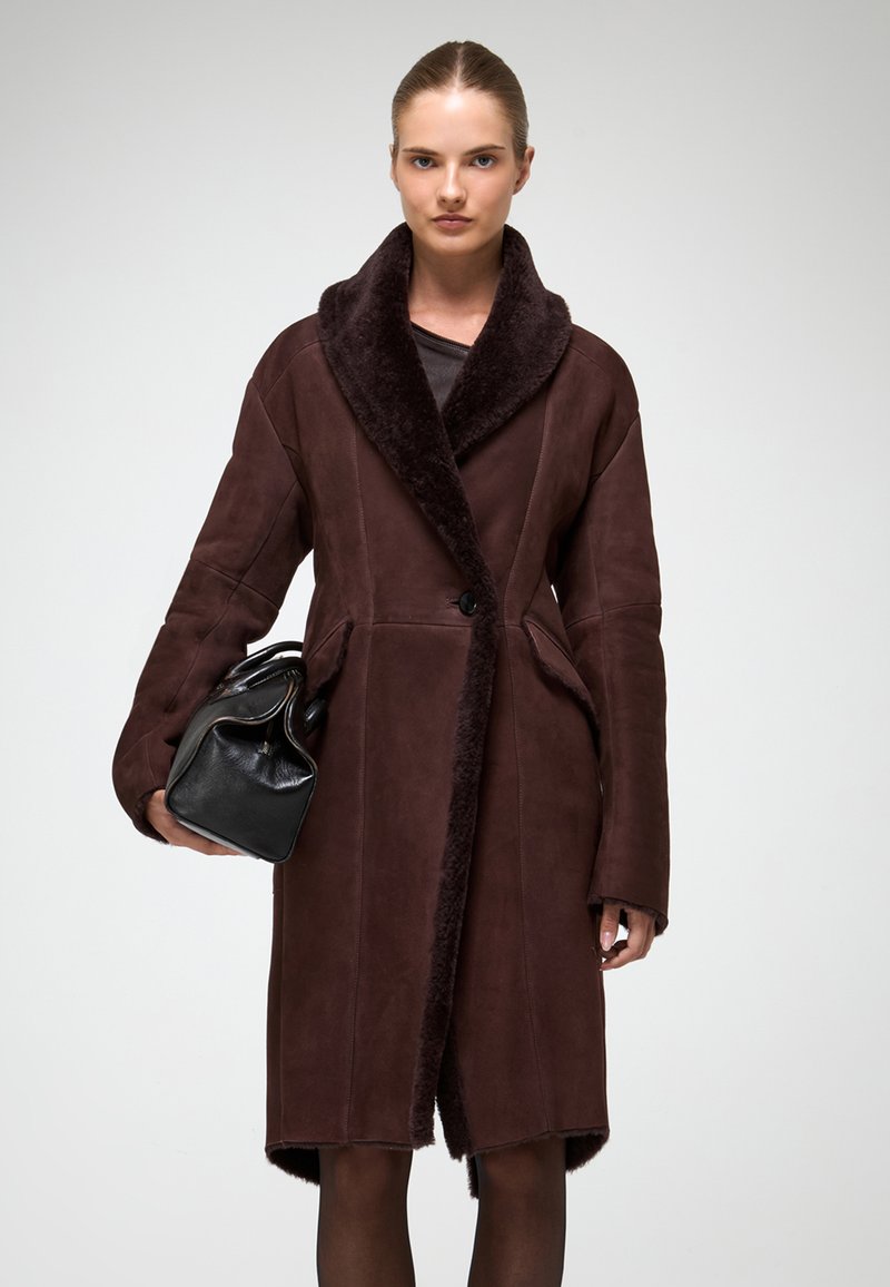 Hnedý kabát z ovčej vlny s širokým šálovým golierom, predným zapínaním na gombíky a bočnými vreckami, nosený cez čierny outfit, doplnený čiernou kabelkou.