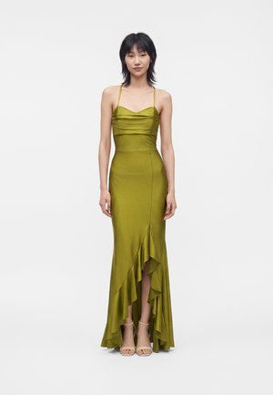Femme portant une robe en satin vert olive sans manches avec des bretelles fines et un ourlet asymétrique à volants, se tenant devant un fond blanc uni.