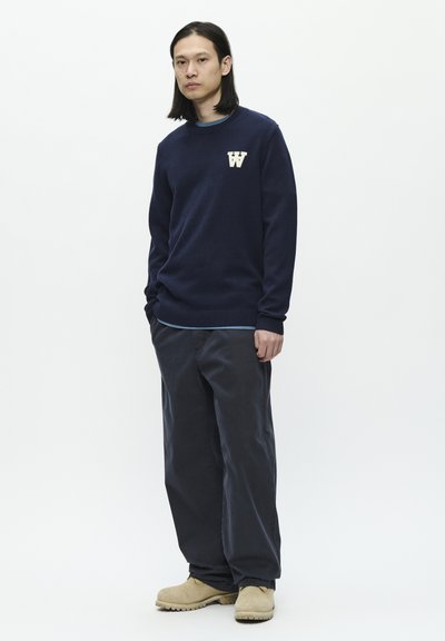 Navy wool sweater with ribbed cuffs and a small white "W" logo, paired with dark grey loose-fit pants and beige clogs.