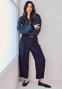 Giacca bomber nera intrecciata con polsini e orlo a costine, abbinata a pantaloni neri larghi e scarpe nere piatte, su uno sfondo di muro chiaro e texture.