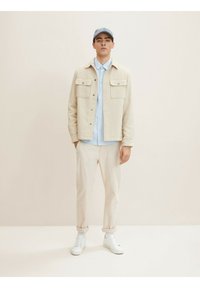 Chaqueta de mezcla de lana beige con dos bolsillos en el pecho, camisa azul clara, pantalones crema y zapatillas blancas. Una gorra gris claro completa el conjunto.