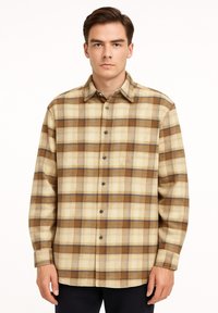 Junger Mann, der ein beiges und braunes kariertes Hemd mit langen Ärmeln trägt und vor einem schlichten weißen Hintergrund steht.