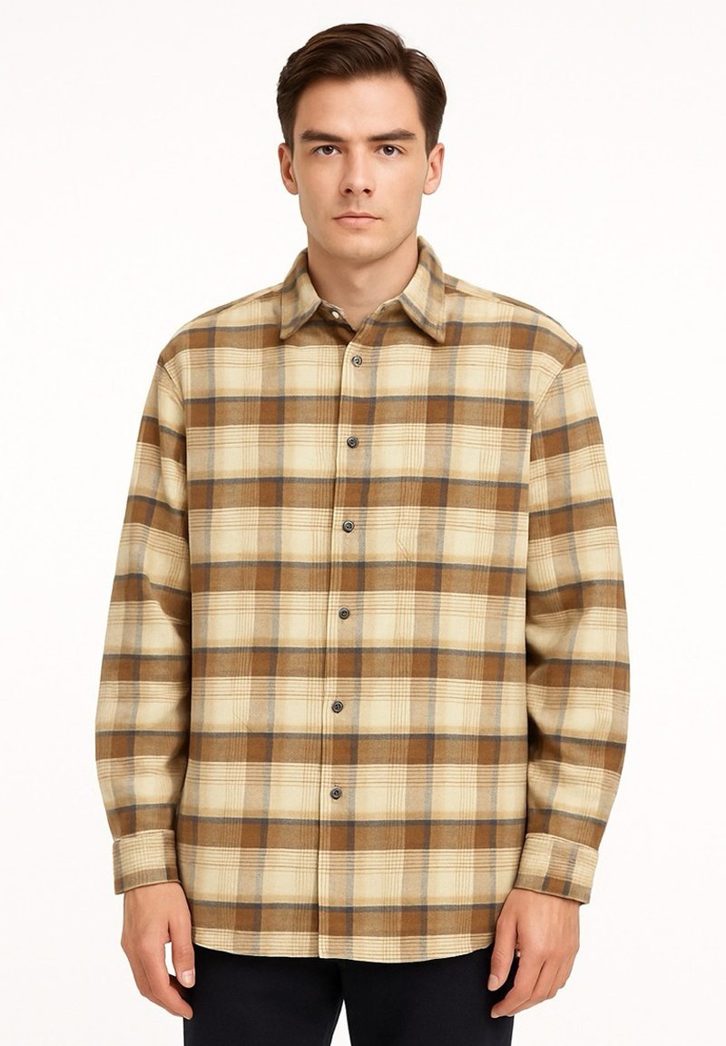 Junger Mann, der ein beiges und braunes kariertes Hemd mit langen Ärmeln trägt und vor einem schlichten weißen Hintergrund steht.