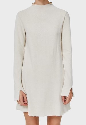 Roomkleurige jurk met lange mouwen, een gerimpelde hoge halslijn en duimgaten in de mouwen, gedragen door een persoon die staat tegen een effen achtergrond.