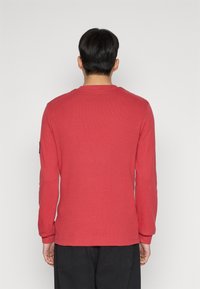 Röd långärmad tröja med textilstruktur, rund hals och ribbmuddar. Har en svart etikett på vänster ärm.