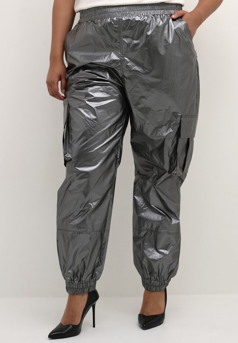 Femme portant un pantalon cargo gris brillant avec des poignets élastiques, associé à des chaussures noires à talons hauts, une main dans la poche.