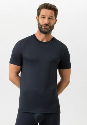 Uomo con barba che indossa una camicia nera aderente a maniche corte e pantaloni neri coordinati, in piedi davanti a uno sfondo bianco semplice.