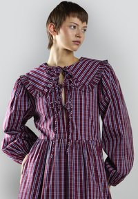 Vestido de cuadros morados con un gran cuello acampanado, escote en forma de ojo de cerradura y detalle de cordón. Mangas largas y voluminosas y falda fruncida. Material de algodón.