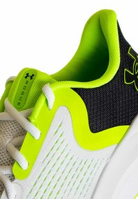 Zapatilla deportiva blanca y negra con llamativos acentos en verde lima, parte superior de malla, acabado texturizado y sistema de cordones. Presenta la marca Under Armour.