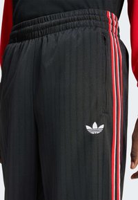 Schwarze Sporthose mit vertikalen Nadelstreifen, roten und weißen Seitenstreifen sowie einem weißen Logo am linken Oberschenkel. Elastischer Bund.