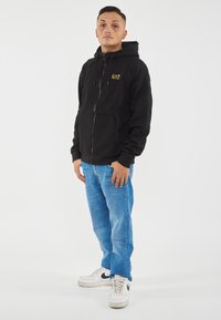 Felpa nera con zip e tasche frontali, con logo dorato. Abbinata a jeans in denim blu e sneakers bianche con dettagli neri.