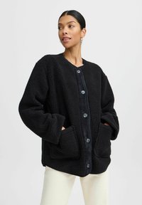 Giacca in pile nera con un esterno texturizzato, scollo tondo, ampie tasche frontali e chiusura con bottoni. Vestibilità morbida e oversize.