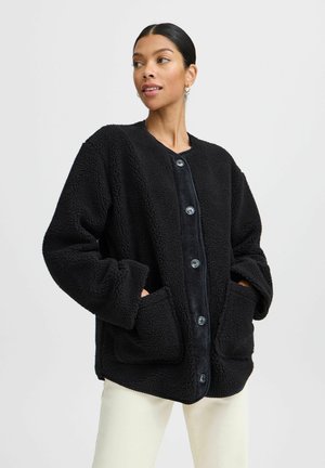 Giacca in pile nera con un esterno texturizzato, scollo tondo, ampie tasche frontali e chiusura con bottoni. Vestibilità morbida e oversize.