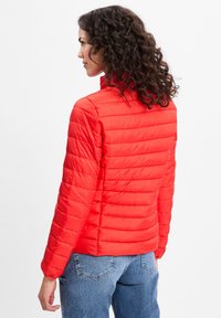 Rode pufferjack met gequiltde textuur, hoge kraag en elastische manchetten. Draagt over blauwe jeans, wat het fitted ontwerp en het lichte materiaal benadrukt.