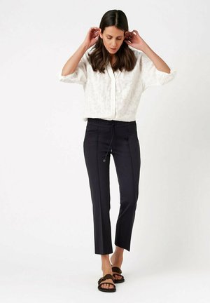 Woman adjusting hair wearing white textured blouse, black slim-fit cropped pants, and dark slip-on sandals, standing against a white background.