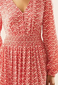 Vestido de flores rojas con mangas largas, escote en V, cintura fruncida y falda fluida. El material parece ligero y texturizado.