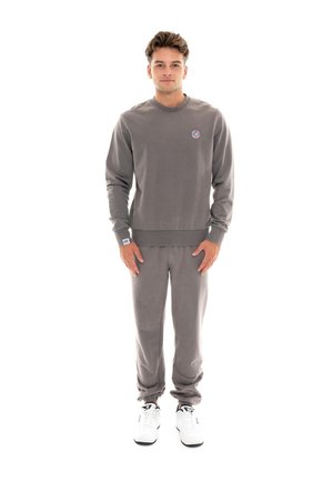 Un homme porte un sweat-shirt gris assorti à un pantalon de jogging et des baskets blanches, sur un fond blanc uni.