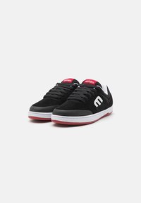 Black suede sneakers with white rubber soles and red accents. Features a padded tongue and a logo on the side. Classic lace-up design.