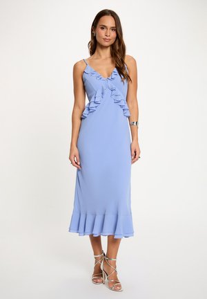 Donna che indossa un vestito midi azzurro chiaro senza maniche con volant e sandali argentati con tacchi alti, in piedi contro uno sfondo bianco neutro.