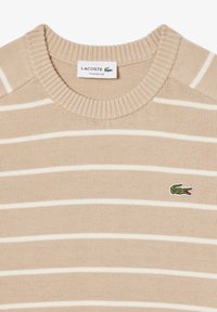 Maglione in cotone beige con strisce orizzontali bianche, scollo a giro e polsini a coste. Presenta il logo Lacoste ricamato in verde.