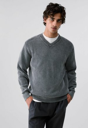 Jeune homme portant un pull gris à côtes en V par-dessus une chemise blanche et un pantalon foncé, debout les mains dans les poches devant un fond uni.