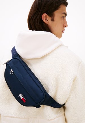 Personne portant une veste polaire blanche et un sac bandoulière Tommy Jeans bleu marine dans le dos, faisant face à droite sur un fond blanc.