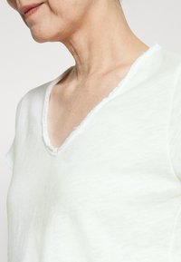 Vaaleanvihreä, lyhythihainen V-pääntiehuppari pehmeästä kankaasta, jossa on karkea reuna kaula-aukossa, ja siinä on hienovarainen rakenne.