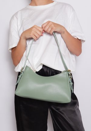 Persona con maglietta bianca e pantaloni neri tiene una borsa in pelle verde chiaro per la cinghia davanti a uno sfondo bianco.