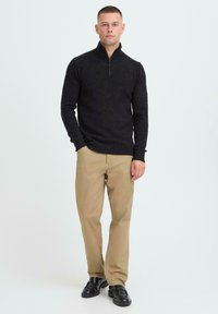 Zwarte zip-neksweater van gestructureerde stof, gecombineerd met beige broek en zwarte schoenen, staande tegen een lichte achtergrond.