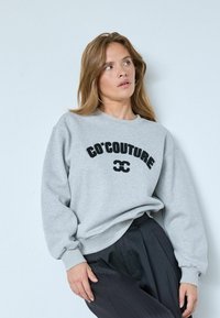 Szürke pulóver fekete "CÔ'COUTURE" hímzéssel, kerek nyakkal és enyhén fodros ujjakkal. Fekete, pliszírozott nadrággal viselve.