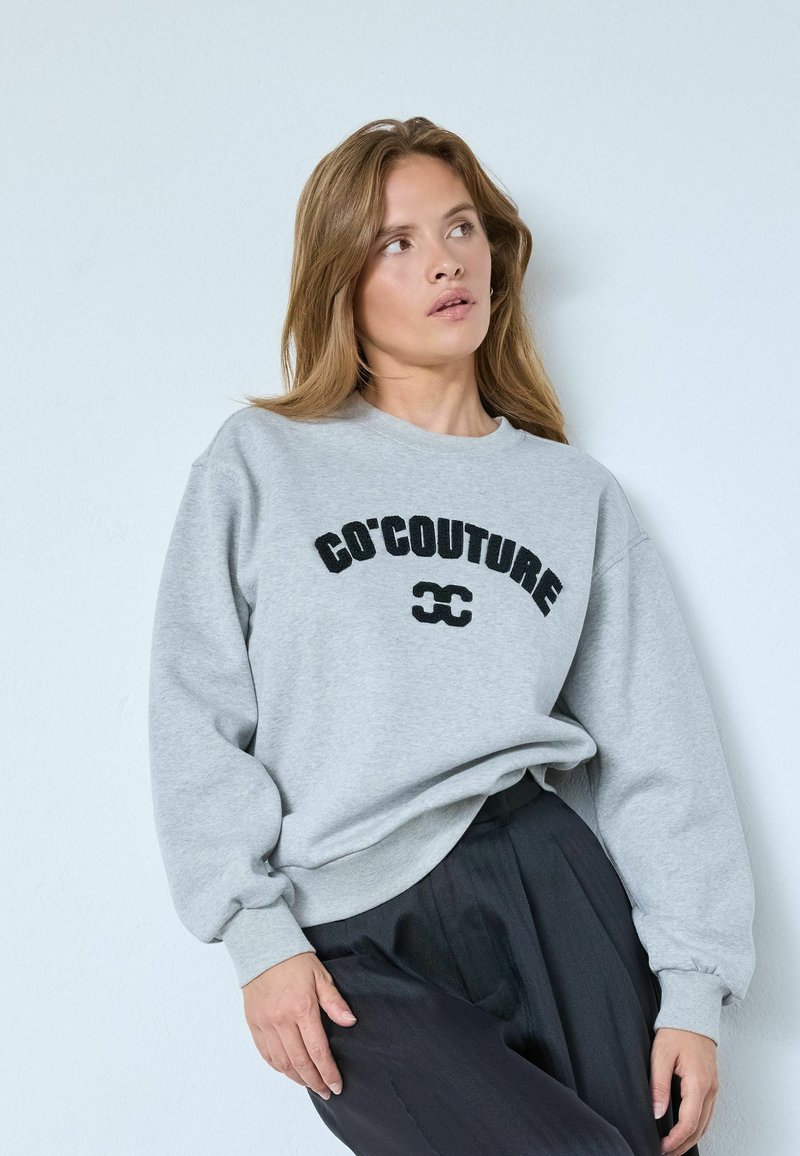 Szürke pulóver fekete "CÔ'COUTURE" hímzéssel, kerek nyakkal és enyhén fodros ujjakkal. Fekete, pliszírozott nadrággal viselve.