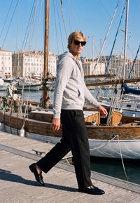 Jeune homme portant des lunettes de soleil, un sweat à capuche gris et un pantalon noir marchant sur un quai à côté de voiliers en bois amarrés dans une marina ensoleillée.