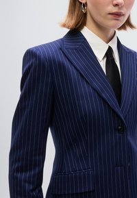 Σκούρο μπλε σακάκι με ρίγες, γιακά με ένωση, ένα κουμπί και τσέπες με καπάκι, φτιαγμένο από λεία ύφασμα. Άσπρο πουκάμισο με γιακά και μαύρη γραβάτα ορατά.