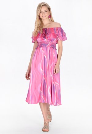 Giovane donna che indossa un vestito con spalle scoperte e volant rosa e viola con sandali, sorridente su uno sfondo bianco.