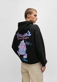 Felpa nera con cappuccio caratterizzata da un design colorato di cartone animato sul retro, raffigurante un personaggio con un hamburger. Materiale morbido, vestibilità ampia, polsini a costine.