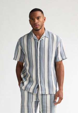 Mann trägt kurzärmeliges, geknöpftes Hemd und passende Hose mit vertikalen blauen, weißen und beigen Streifen vor einfarbigem Hintergrund.