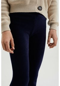 Colanți cu nervuri de culoare închisă, fabricați dintr-un material moale, cu croi mulat și model texturat. O jachetă cardigan de tricot, de culoare crem, cu un buton, arătând parțial.