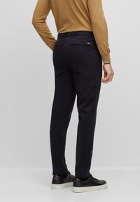 Muž v černých slim-fit kalhotách, černých teniskách a hnědém dlouhém rukávu, stojící na jednobarevném šedém pozadí.