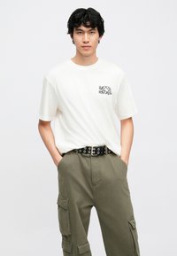 Hvid t-shirt med teksten "HAPPY HOLIDAYS" trykt i sort på brystet, kombineret med olivengrønne cargobukser og et sort bælte med nitter.