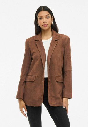 Woman wearing a brown suede blazer over a white top and black pants, standing against a plain white background.