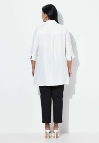 White long-sleeve top with back pleat detail, paired with black trousers. The outfit features tailored fit and open cuffs. White high-heeled shoes.