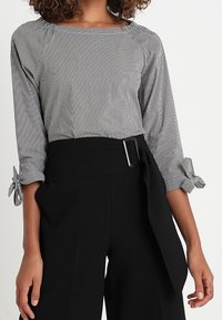 Maglietta a righe bianco e nero con maniche a tre quarti e dettaglio a nodo sui polsini, abbinata a pantaloni larghi neri a vita alta con cintura.