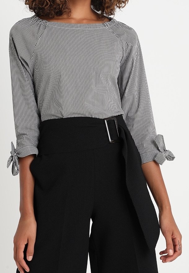Maglietta a righe bianco e nero con maniche a tre quarti e dettaglio a nodo sui polsini, abbinata a pantaloni larghi neri a vita alta con cintura.