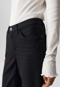 Jeans in denim nero presentano una vestibilità slim, tasche frontali e una chiusura con bottone. Una camicia bianca a maniche lunghe ha polsini rifiniti a onda.