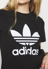 Must must lahe must t-särk, millel on suur valge Adidas'i logo koos kolme triibu ja kolmiku graafikaga rinna peal.