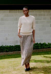 Beige, flauschiger Strickpullover mit kurzen Ärmeln, kombiniert mit einem schwarz-weiß gestreiften, fließenden Maxikleid. Das Modell steht in einem grasbewachsenen Bereich.