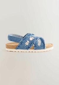 Sandal in denim con cinturini azzurri sfrangiati, ricamo floreale e suola in sughero. Presenta una suola in gomma bianca con motivo a onde.