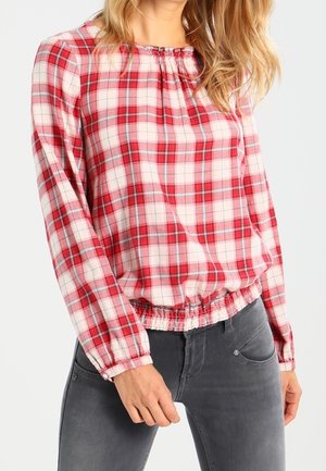 Vrouw draagt een rood-wit geruit blouse met lange mouwen, met een geplooide halslijn en zoom, gecombineerd met donkergrijze spijkerbroek.
